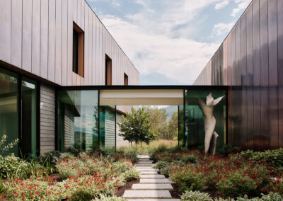 A modern residence with copper cladding and glass walls, featuring a central garden filled with greenery and red flowers, and a white abstract sculpture. The building's clean lines and natural materials harmonize with the surrounding landscape under a partly cloudy sky