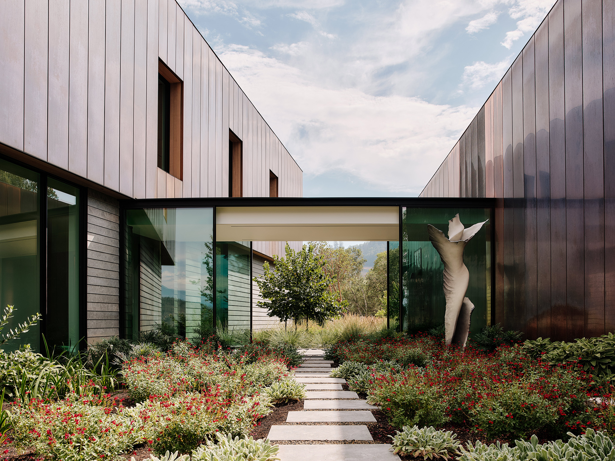 A modern residence with copper cladding and glass walls, featuring a central garden filled with greenery and red flowers, and a white abstract sculpture. The building's clean lines and natural materials harmonize with the surrounding landscape under a partly cloudy sky