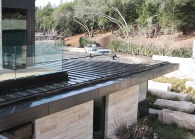 A modern residence with copper cladding and glass walls, featuring a central garden filled with greenery and red flowers, and a white abstract sculpture. The building's clean lines and natural materials harmonize with the surrounding landscape under a partly cloudy sky